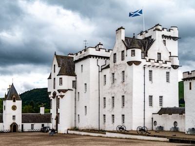 A historic white castle with multiple towers and crenellated parapets stands under a cloudy sky. The building features a Scottish flag on a pole, dark roof tiles, and small windows. Black cannons are lined up on the gravel courtyard alongside the building. Two people are visible near the entrance of a smaller tower with a clock.