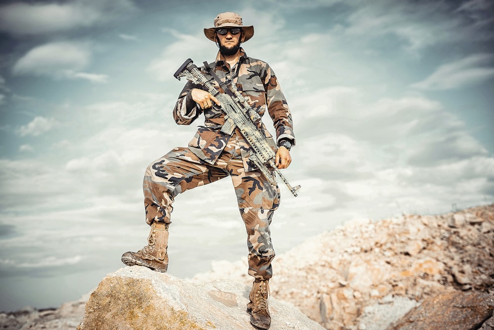 hombre vestido con uniforme de camuflaje marrón y verde sosteniendo un rifle mientras está de pie sobre una roca