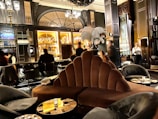 A luxurious lounge with elegant, plush seating and a sophisticated bar. The interior features dark wood paneling, ornate lighting fixtures, and a stunning backdrop of illuminated bottles on shelves. The atmosphere is warm and inviting, with various patrons sitting at the bar and others moving around the space. Large decorative plumes and an opulent chandelier add to the grandeur of the room.