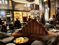 A luxurious lounge with elegant, plush seating and a sophisticated bar. The interior features dark wood paneling, ornate lighting fixtures, and a stunning backdrop of illuminated bottles on shelves. The atmosphere is warm and inviting, with various patrons sitting at the bar and others moving around the space. Large decorative plumes and an opulent chandelier add to the grandeur of the room.