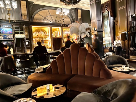 A luxurious lounge with elegant, plush seating and a sophisticated bar. The interior features dark wood paneling, ornate lighting fixtures, and a stunning backdrop of illuminated bottles on shelves. The atmosphere is warm and inviting, with various patrons sitting at the bar and others moving around the space. Large decorative plumes and an opulent chandelier add to the grandeur of the room.