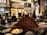 A luxurious lounge with elegant, plush seating and a sophisticated bar. The interior features dark wood paneling, ornate lighting fixtures, and a stunning backdrop of illuminated bottles on shelves. The atmosphere is warm and inviting, with various patrons sitting at the bar and others moving around the space. Large decorative plumes and an opulent chandelier add to the grandeur of the room.