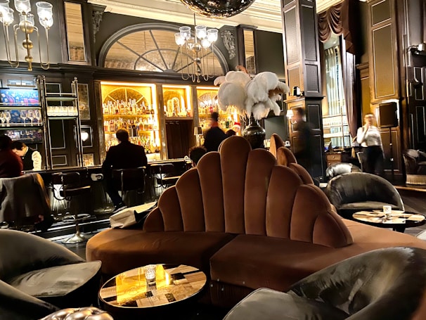 A luxurious lounge with elegant, plush seating and a sophisticated bar. The interior features dark wood paneling, ornate lighting fixtures, and a stunning backdrop of illuminated bottles on shelves. The atmosphere is warm and inviting, with various patrons sitting at the bar and others moving around the space. Large decorative plumes and an opulent chandelier add to the grandeur of the room.