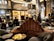 A luxurious lounge with elegant, plush seating and a sophisticated bar. The interior features dark wood paneling, ornate lighting fixtures, and a stunning backdrop of illuminated bottles on shelves. The atmosphere is warm and inviting, with various patrons sitting at the bar and others moving around the space. Large decorative plumes and an opulent chandelier add to the grandeur of the room.