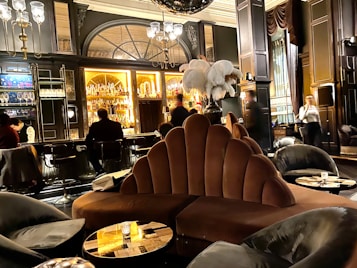A luxurious lounge with elegant, plush seating and a sophisticated bar. The interior features dark wood paneling, ornate lighting fixtures, and a stunning backdrop of illuminated bottles on shelves. The atmosphere is warm and inviting, with various patrons sitting at the bar and others moving around the space. Large decorative plumes and an opulent chandelier add to the grandeur of the room.