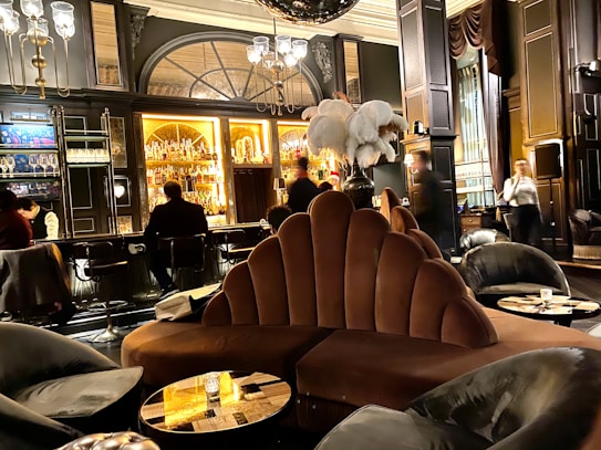 A luxurious lounge with elegant, plush seating and a sophisticated bar. The interior features dark wood paneling, ornate lighting fixtures, and a stunning backdrop of illuminated bottles on shelves. The atmosphere is warm and inviting, with various patrons sitting at the bar and others moving around the space. Large decorative plumes and an opulent chandelier add to the grandeur of the room.