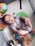 A baby lies on a playmat, wearing a printed onesie. The baby holds its feet up playfully, surrounded by colorful toys including a multicolored ball and a soft book with animal illustrations. The playmat has a textured, patterned surface with printed images like a ladybug and a leaf.