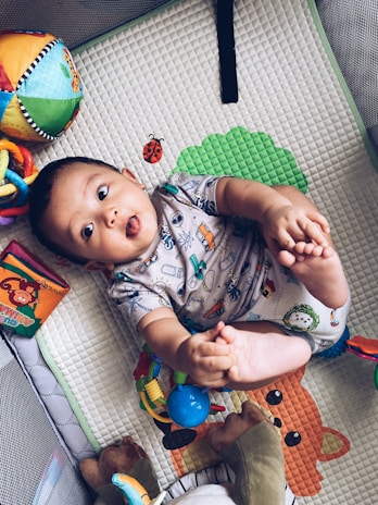 A baby lies on a playmat, wearing a printed onesie. The baby holds its feet up playfully, surrounded by colorful toys including a multicolored ball and a soft book with animal illustrations. The playmat has a textured, patterned surface with printed images like a ladybug and a leaf.