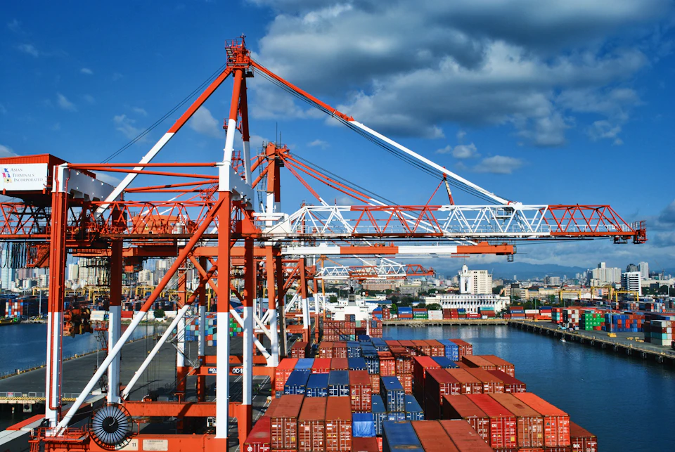 The image depicts a bustling port scene with a large red and white crane towering over the water, lifting a blue and white cargo ship into position on the water. The crane is surrounded by several other cranes and ships, indicating a busy and active port environment. In the background, there are buildings of various sizes, suggesting that this port is located in an urban area with commercial or industrial facilities nearby.