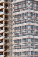 A partially completed building covered in scaffolding features prominently. The structure shows layers of concrete and metal with a grid of beams and platforms, highlighting the construction phase. The facade is enveloped in protective netting, while partially open sections reveal stacks of building materials.