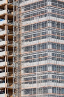 A partially completed building covered in scaffolding features prominently. The structure shows layers of concrete and metal with a grid of beams and platforms, highlighting the construction phase. The facade is enveloped in protective netting, while partially open sections reveal stacks of building materials.