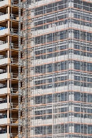 A partially completed building covered in scaffolding features prominently. The structure shows layers of concrete and metal with a grid of beams and platforms, highlighting the construction phase. The facade is enveloped in protective netting, while partially open sections reveal stacks of building materials.