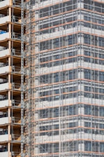 A partially completed building covered in scaffolding features prominently. The structure shows layers of concrete and metal with a grid of beams and platforms, highlighting the construction phase. The facade is enveloped in protective netting, while partially open sections reveal stacks of building materials.