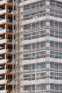 A partially completed building covered in scaffolding features prominently. The structure shows layers of concrete and metal with a grid of beams and platforms, highlighting the construction phase. The facade is enveloped in protective netting, while partially open sections reveal stacks of building materials.