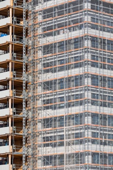 A partially completed building covered in scaffolding features prominently. The structure shows layers of concrete and metal with a grid of beams and platforms, highlighting the construction phase. The facade is enveloped in protective netting, while partially open sections reveal stacks of building materials.