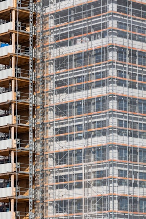 A partially completed building covered in scaffolding features prominently. The structure shows layers of concrete and metal with a grid of beams and platforms, highlighting the construction phase. The facade is enveloped in protective netting, while partially open sections reveal stacks of building materials.