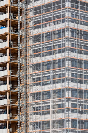 A partially completed building covered in scaffolding features prominently. The structure shows layers of concrete and metal with a grid of beams and platforms, highlighting the construction phase. The facade is enveloped in protective netting, while partially open sections reveal stacks of building materials.