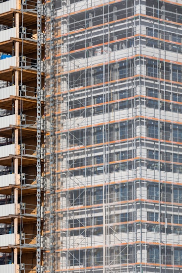 A partially completed building covered in scaffolding features prominently. The structure shows layers of concrete and metal with a grid of beams and platforms, highlighting the construction phase. The facade is enveloped in protective netting, while partially open sections reveal stacks of building materials.