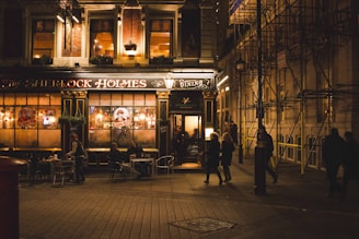 An inviting pub exterior warmly lit at night, with people sitting at tables outside. The establishment has a classic and sophisticated design with large windows and a black and gold color scheme. Signs and string lights add charm to the scene. Several pedestrians pass by on the cobblestone street.