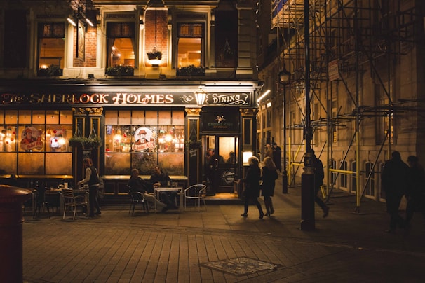 An inviting pub exterior warmly lit at night, with people sitting at tables outside. The establishment has a classic and sophisticated design with large windows and a black and gold color scheme. Signs and string lights add charm to the scene. Several pedestrians pass by on the cobblestone street.