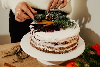 A person is decorating a cake with white frosting and berry sauce drizzled on top. The cake is adorned with pine branches, cinnamon sticks, and a small pine cone. A pair of hands is carefully placing the decorations. The scene includes a wooden surface with a bow and festive greenery, creating a rustic, holiday atmosphere.