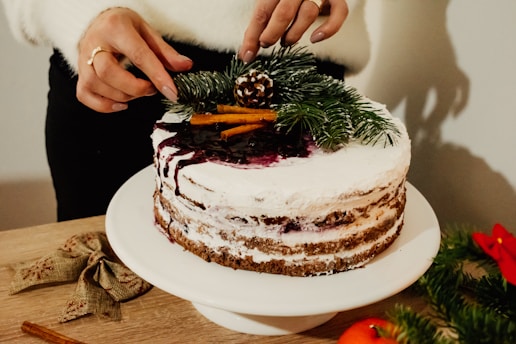 A person is decorating a cake with white frosting and berry sauce drizzled on top. The cake is adorned with pine branches, cinnamon sticks, and a small pine cone. A pair of hands is carefully placing the decorations. The scene includes a wooden surface with a bow and festive greenery, creating a rustic, holiday atmosphere.