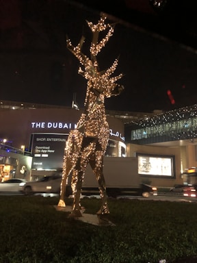 A metallic sculpture of a reindeer adorned with twinkling lights stands in front of a well-lit building during the night. The structure is surrounded by greenery, with vehicles passing by on the road nearby. The building features illuminated signage, suggesting a shopping or entertainment venue.