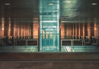 Elevated view of a modern, symmetrical architectural structure featuring a glass elevator in the center surrounded by reflective metal surfaces. The setting is illuminated with a warm, ambient light, casting subtle reflections and shadows.