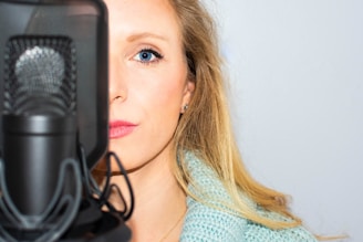 A woman with blonde hair, partially obscured by a large microphone, appears focused. She is wearing a light blue knitted top against a plain white background.