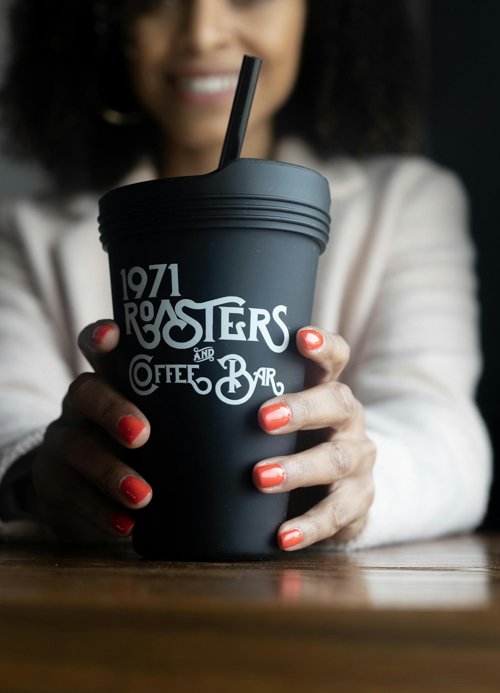 woman wearing white jacket sitting while holding black and white 1971 Roasters and Coffee Bar coffee cup with lid and straw