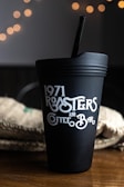 A black coffee tumbler with the text '1971 Roasters and Coffee Bar' in decorative white lettering is situated on a wooden surface. In the background, there is a blurred appearance of warm, glowing bokeh lights and a textured fabric, possibly a burlap sack.
