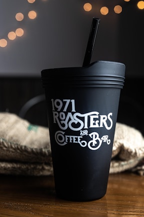 A black coffee tumbler with the text '1971 Roasters and Coffee Bar' in decorative white lettering is situated on a wooden surface. In the background, there is a blurred appearance of warm, glowing bokeh lights and a textured fabric, possibly a burlap sack.