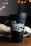 A black coffee tumbler with the text '1971 Roasters and Coffee Bar' in decorative white lettering is situated on a wooden surface. In the background, there is a blurred appearance of warm, glowing bokeh lights and a textured fabric, possibly a burlap sack.