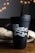 A black coffee tumbler with the text '1971 Roasters and Coffee Bar' in decorative white lettering is situated on a wooden surface. In the background, there is a blurred appearance of warm, glowing bokeh lights and a textured fabric, possibly a burlap sack.