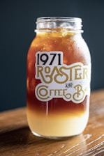 A mason jar sits on a wooden surface, filled with a layered iced coffee drink. The top layer is a dark brown, possibly cold brew or iced coffee, and the bottom layer is a creamy light beige, suggesting milk or cream. The jar is labeled with '1971 Roaster and Coffee Bar' in stylized typography. The background is a dark teal color, providing contrast to the jar and its contents.