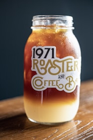 A mason jar sits on a wooden surface, filled with a layered iced coffee drink. The top layer is a dark brown, possibly cold brew or iced coffee, and the bottom layer is a creamy light beige, suggesting milk or cream. The jar is labeled with '1971 Roaster and Coffee Bar' in stylized typography. The background is a dark teal color, providing contrast to the jar and its contents.