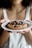 A person is holding a white plate with a freshly baked chocolate chip cookie. The cookie is large, thick, and has generous chunks of chocolate. The person's nails are painted red, and they are wearing a light-colored tank top.