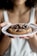 A person is holding a white plate with a freshly baked chocolate chip cookie. The cookie is large, thick, and has generous chunks of chocolate. The person's nails are painted red, and they are wearing a light-colored tank top.