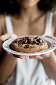 A person is holding a white plate with a freshly baked chocolate chip cookie. The cookie is large, thick, and has generous chunks of chocolate. The person's nails are painted red, and they are wearing a light-colored tank top.