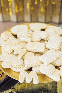 A platter of white iced cookies in various shapes, such as hearts, rectangles, and wavy letters. The cookies are decorated with small silver edible beads, and the platter is placed on a reflective golden surface with a blurred golden backdrop.