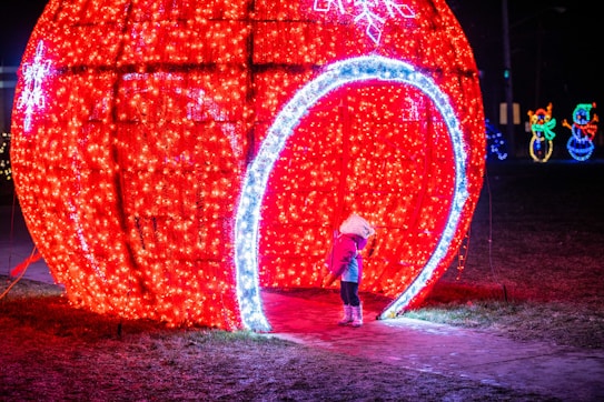 A child in warm winter clothing stands at the entrance of a large, illuminated red Christmas ornament display. The ornament is adorned with bright lights, and there are additional decorations in the background, including festive figures made from lights.