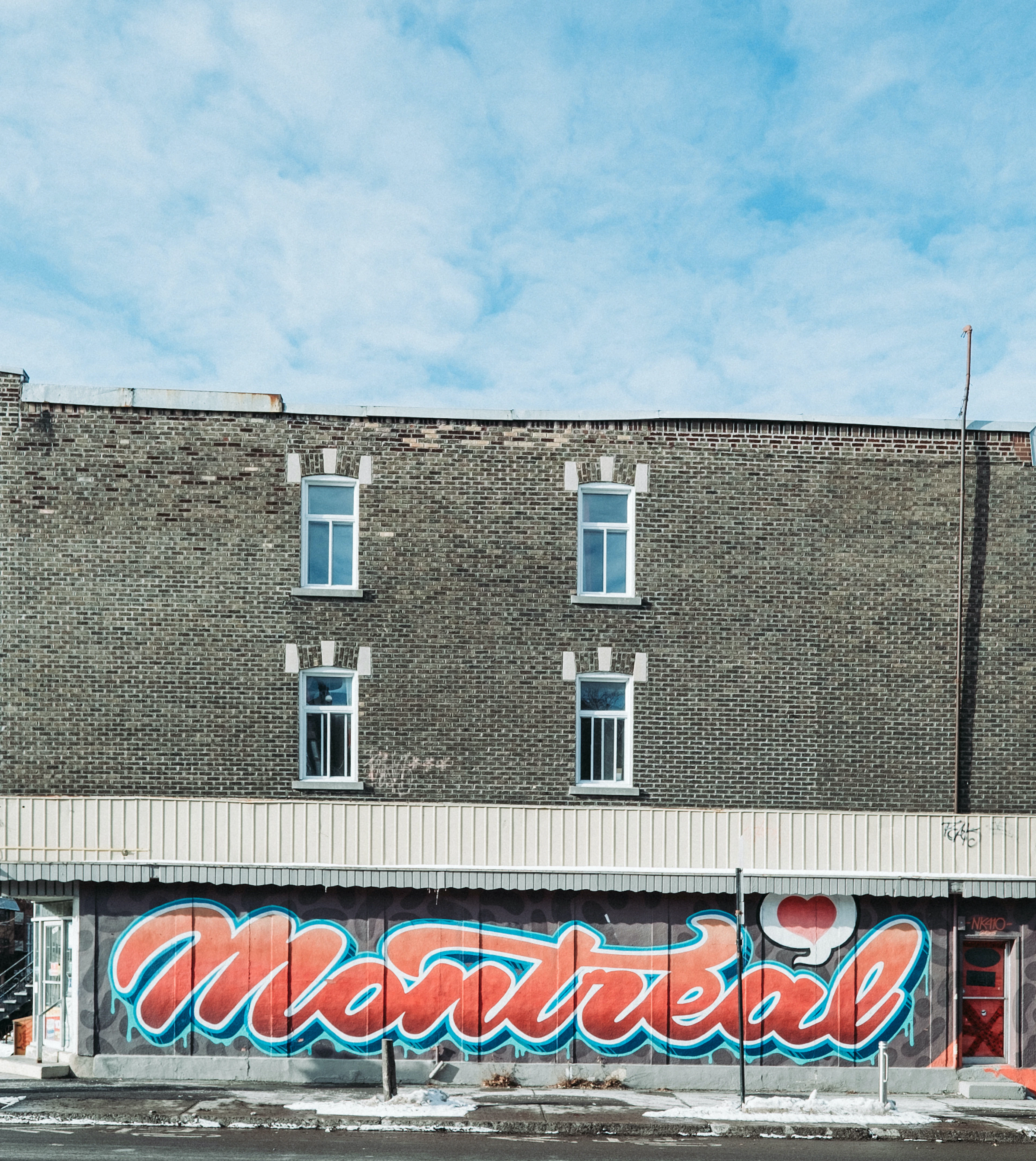 A cursive Montreal mural in red and teal covers a storefront on a brick building under a blue sky.