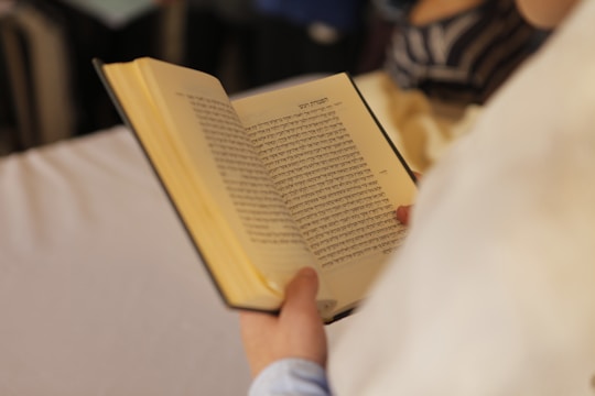 A person is holding an open book with text written in Hebrew. The focus is on the book, with blurred background details.