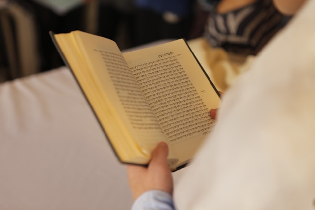 A person is holding an open book with text written in Hebrew. The focus is on the book, with blurred background details.