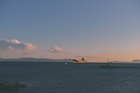 A large cargo ship sails under a clear sky at sunset, with scattered clouds reflecting a warm orange glow. The ship is positioned on a vast body of water with gentle waves, and a small jetty with a lighthouse is visible in the foreground. Distant mountains form a faint horizon line across the sea.
