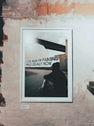 A framed photograph is displayed on a textured, partially exposed brick wall. The photograph features a silhouette of a person sitting on the ground, facing a modern bridge or industrial structure. Text on the image reads, 'Look Mum I'm painting walls legally now.' The setting suggests an urban environment.