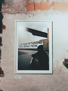 A framed photograph is displayed on a textured, partially exposed brick wall. The photograph features a silhouette of a person sitting on the ground, facing a modern bridge or industrial structure. Text on the image reads, 'Look Mum I'm painting walls legally now.' The setting suggests an urban environment.
