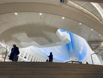 A spacious, modern architectural interior featuring curved, white ceilings and walls with integrated lighting. In the background, a large window reveals a bright, blue sky. Several people are walking up a wide staircase in the foreground, suggesting movement and usage of the space.