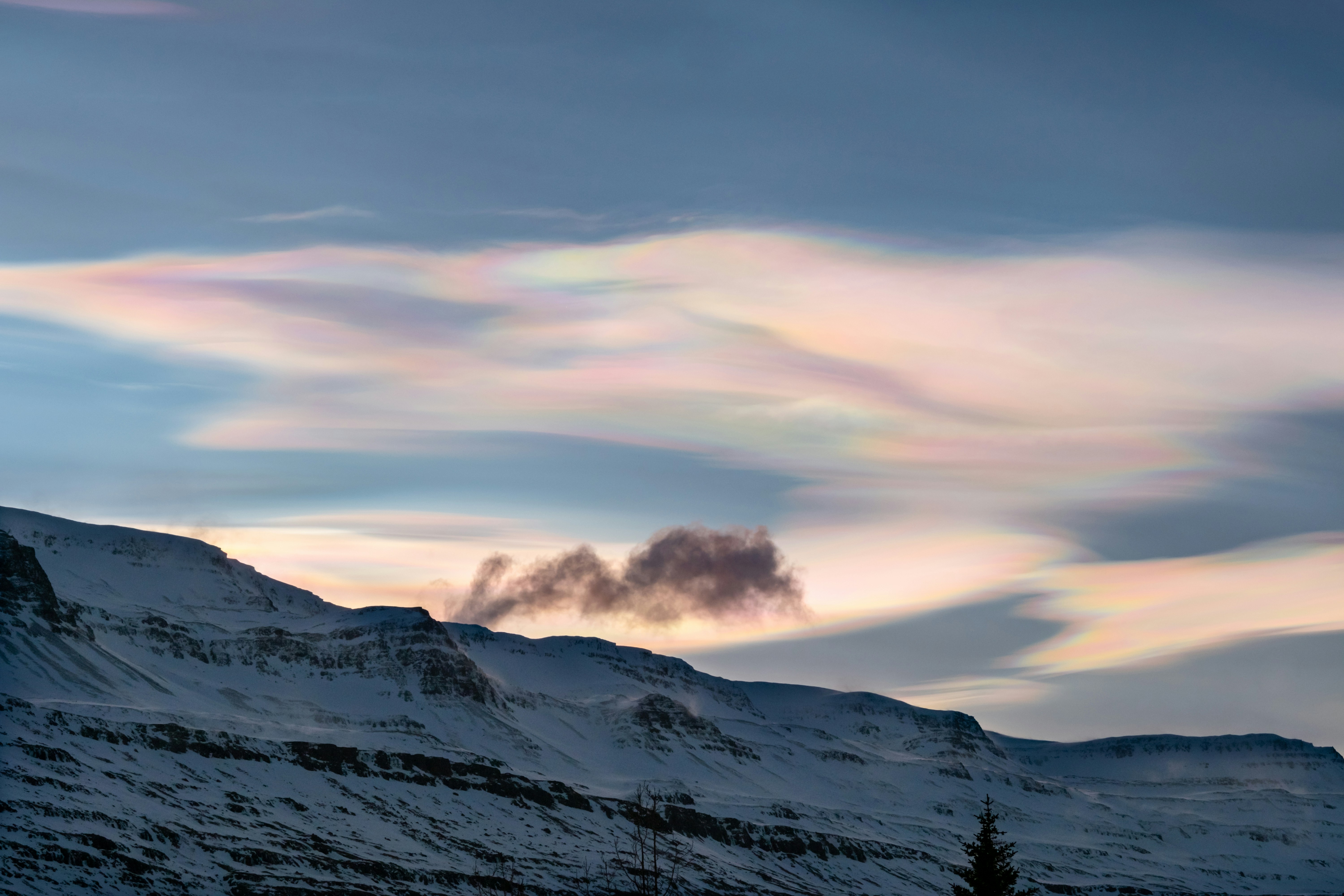 a mountain covered in snow under a cloudy sky, Iridescent clouds above Eastern Iceland.