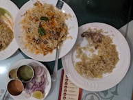 A spread of Indian cuisine with a large serving of biryani garnished with herbs on a white plate, alongside another plate with a partially eaten piece of chicken and rice. There is also a plate containing sliced onions and lemon, with three metal containers holding different sauces or chutneys.